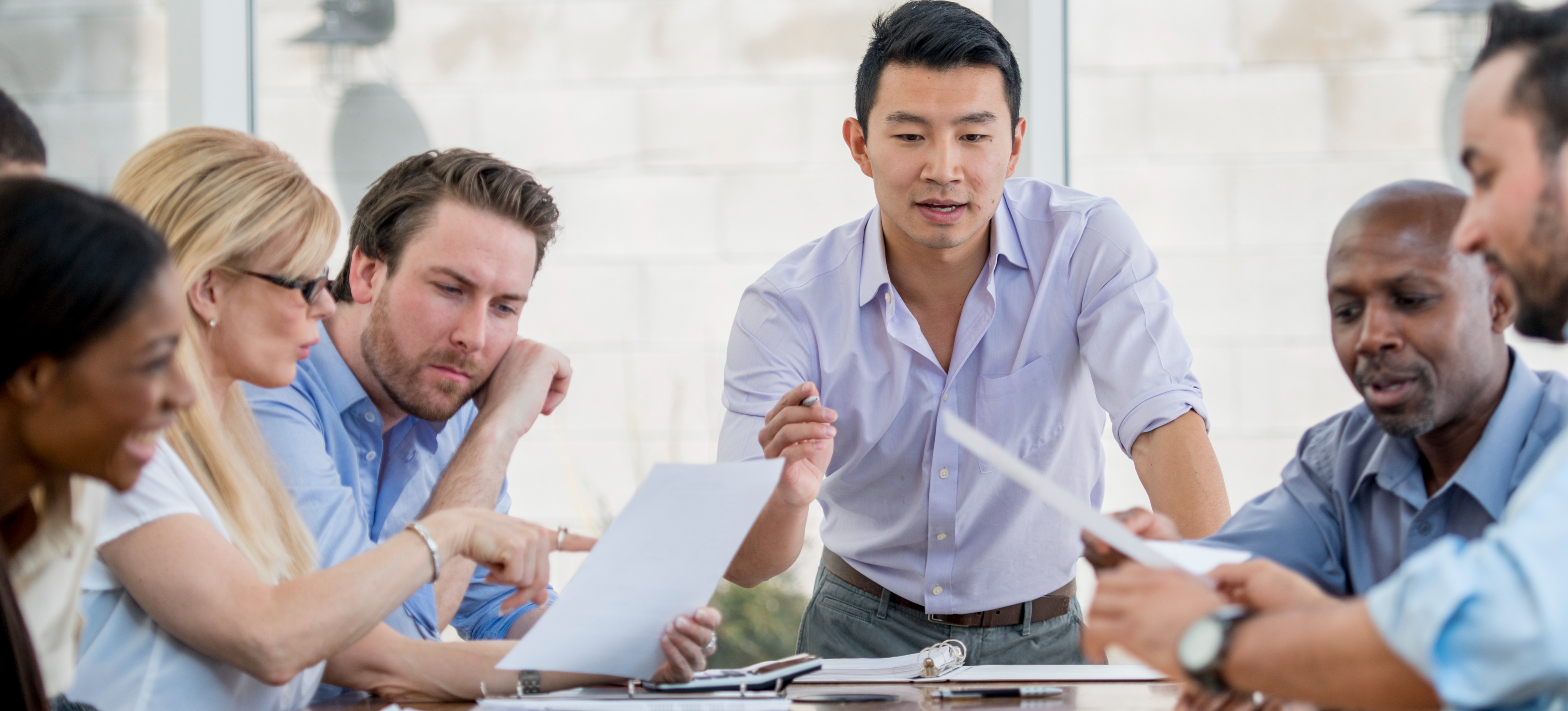 [Featured Image] A team leader stands at a conference table, discussing the business process with five other colleagues, who are passing a paper around.
