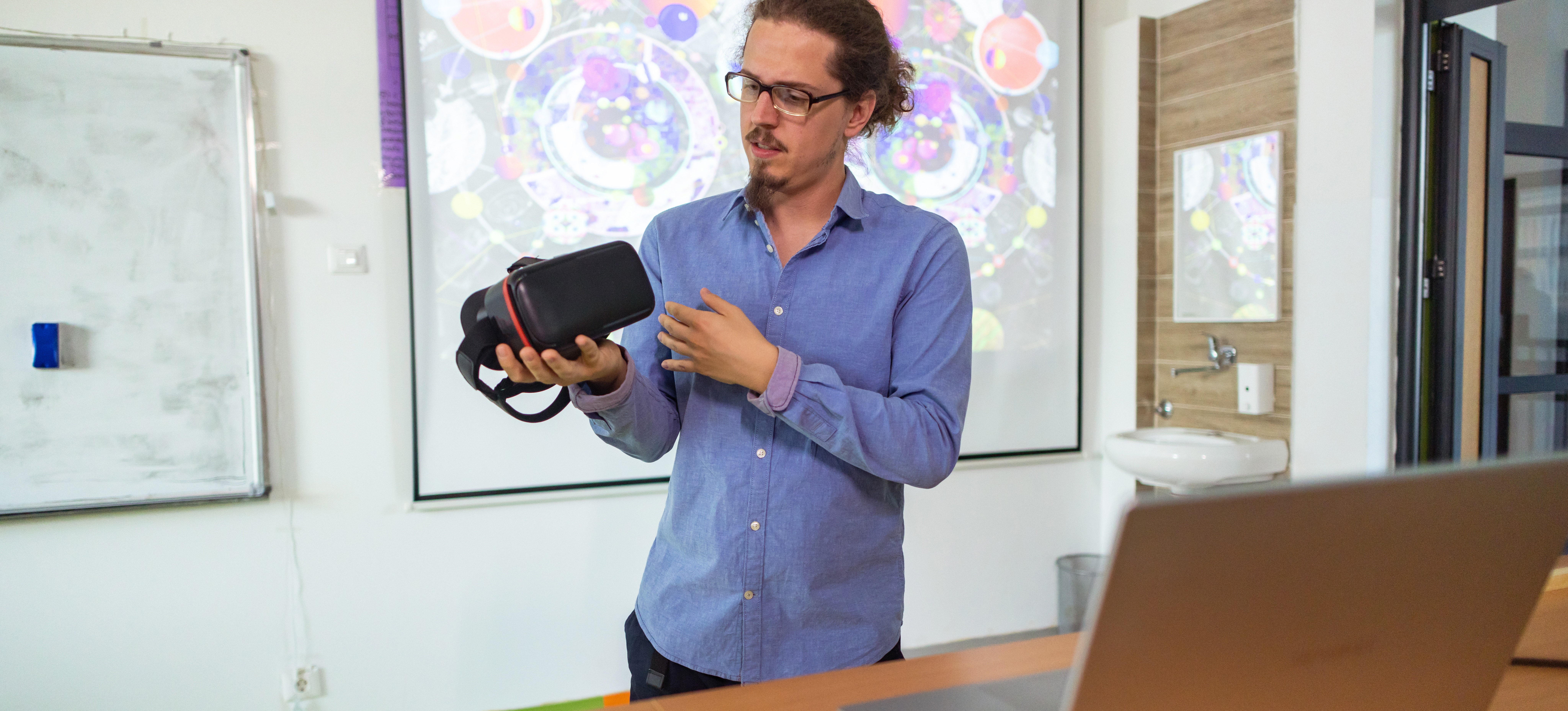 [Image en vedette] Un professeur se tient devant une salle de classe avec un casque de réalité virtuelle pour expliquer aux étudiants le fonctionnement de la réalité virtuelle.