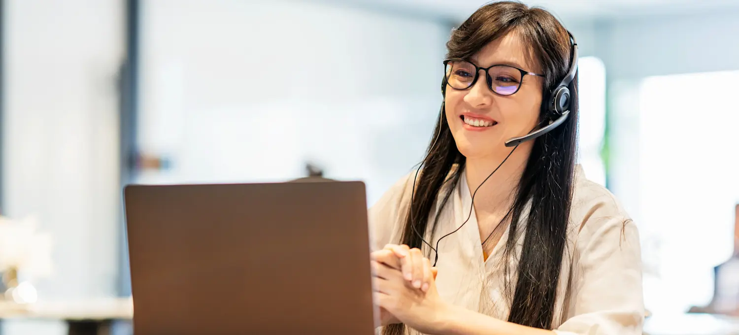 [Featured Image] Working on a sales representative salary, a smiling person wearing a headset talks to a customer remotely over a Zoom meeting on their laptop.
