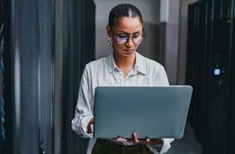 [Featured Image] A woman works on a laptop computer in a server room.