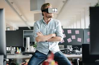 [Featured image] A person in a blue shirt leans against a desk with their arms crossed while wearing a white VR headset.
