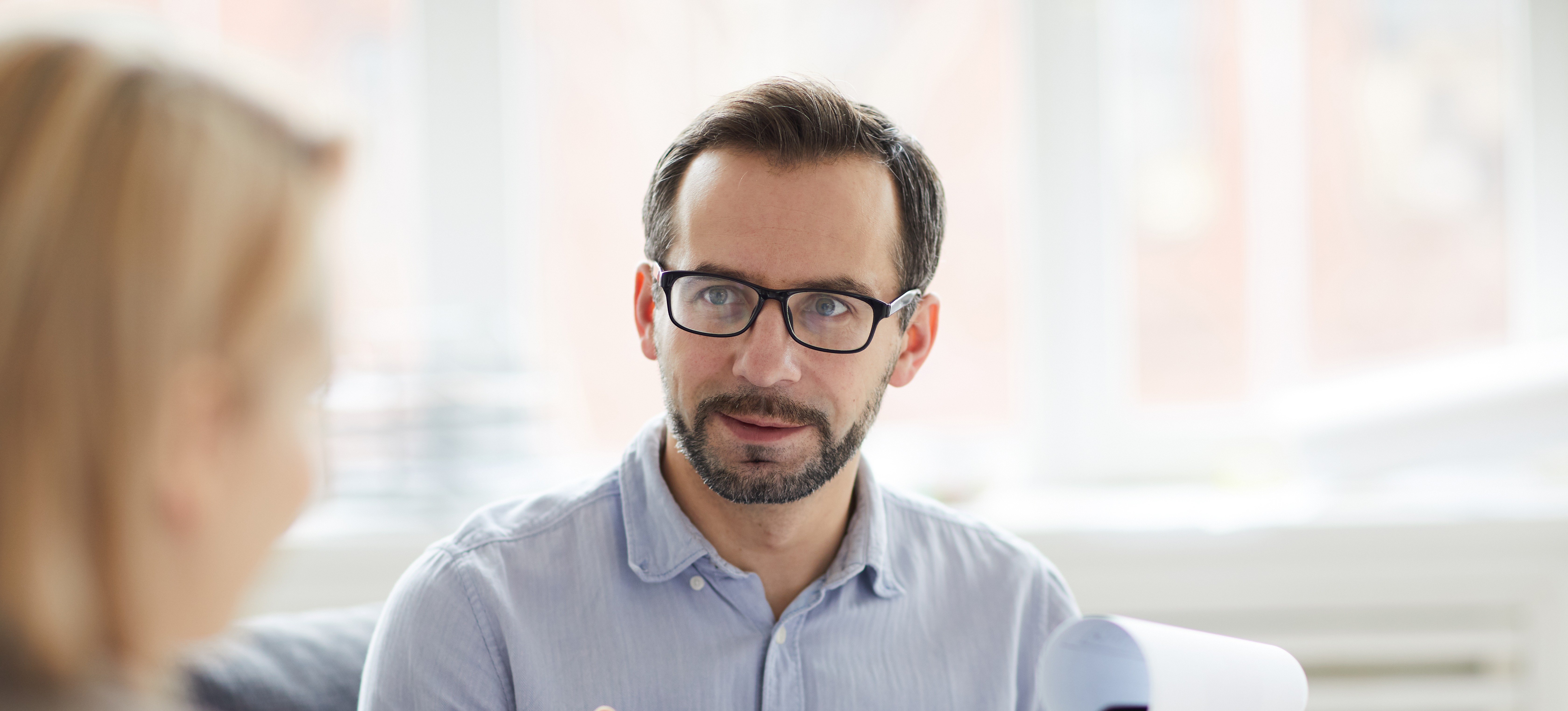 [Featured Image] A genetic counselor, wearing a blue shirt and glasses, is consulting with a patient with blonde hair.        