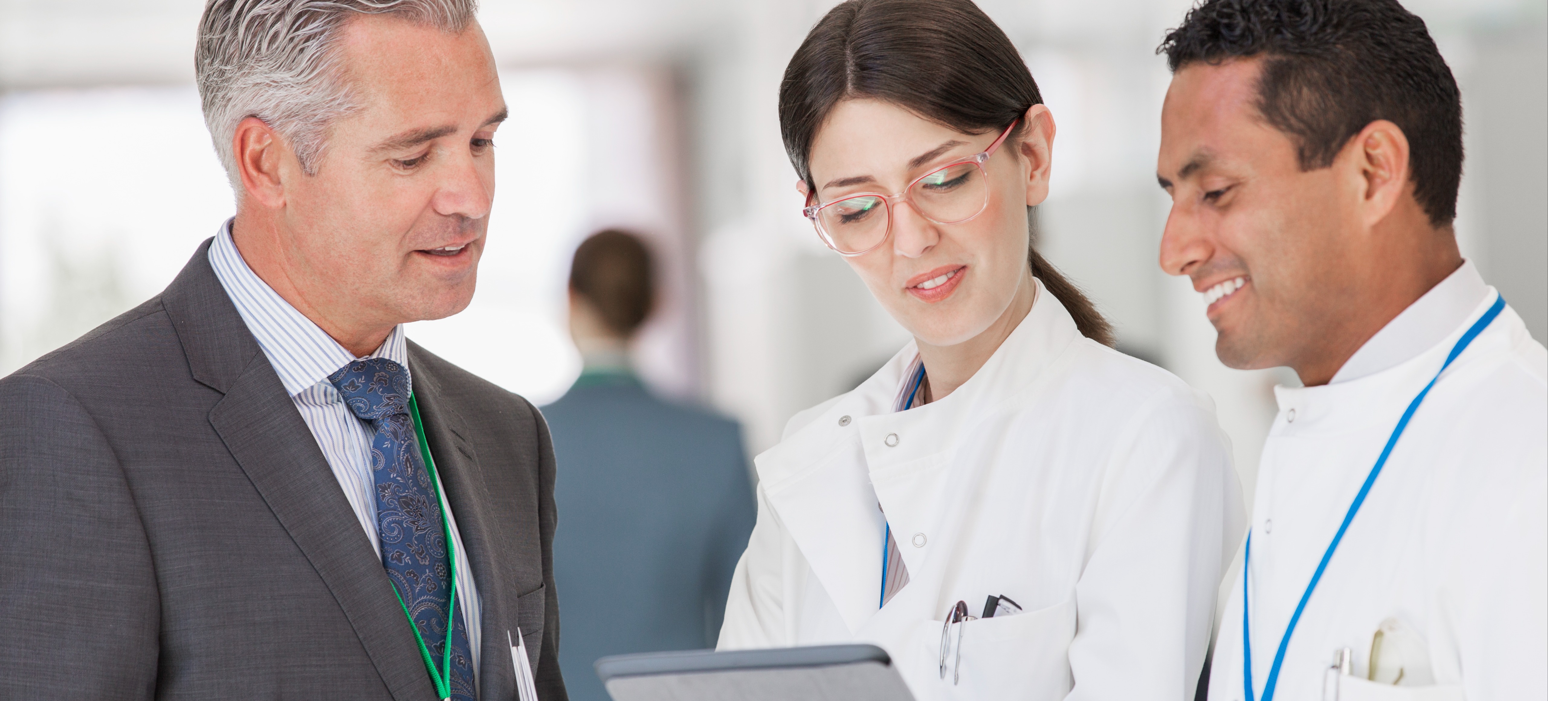 [Featured Image] Three professional health administration degree holders wearing badges, business suits, and white coats converse in a hospital hallway while looking at information on a tablet.
