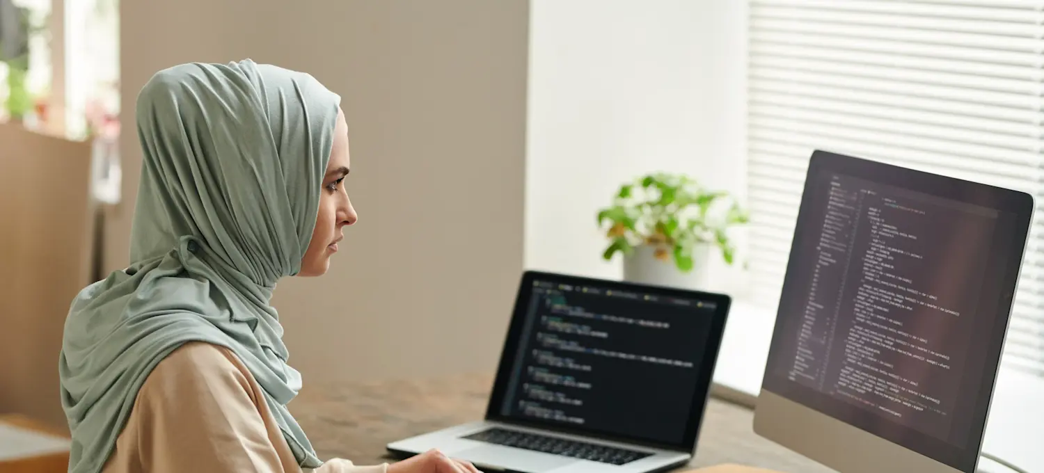 [Featured Image]: A person wearing a tan blouse and a head covering is sitting in front of two computer screens. 