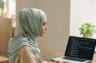 [Featured Image]: A woman wearing a tan blouse and a head covering. She is sitting in front of two computer screens. There is a white headset on the desk. 