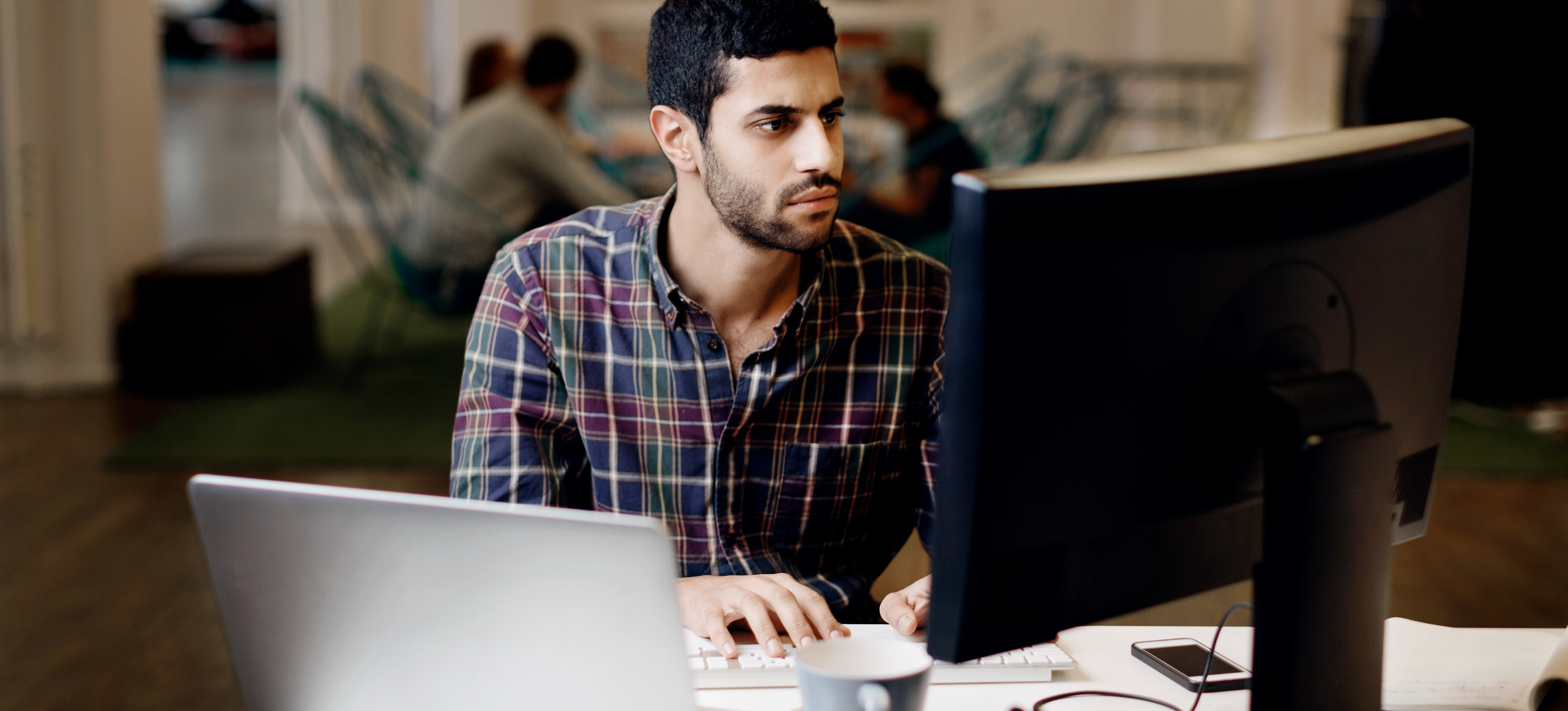 [Featured Image] A person in a software engineering role works at their computer and laptop while their colleagues have a meeting around a table in the background.
