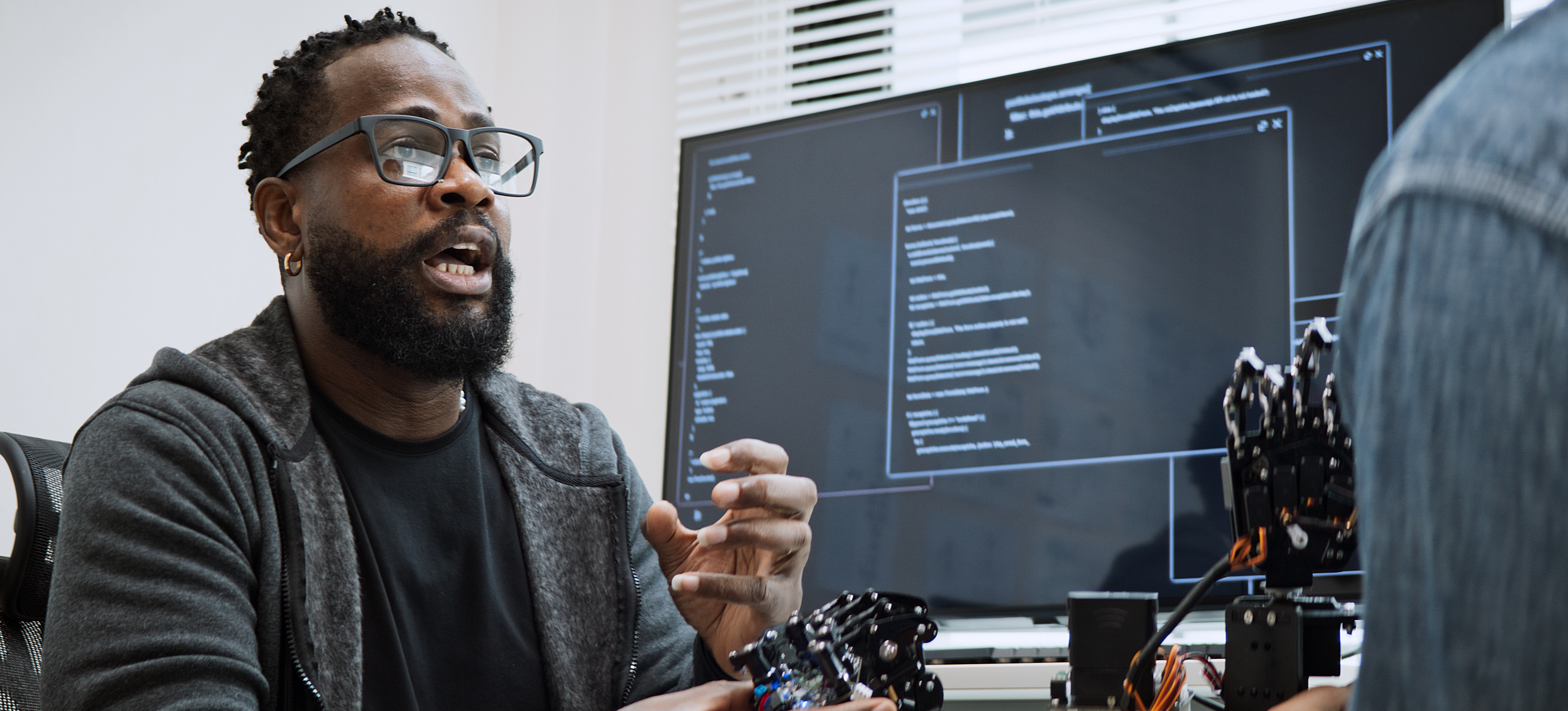 [Featured Image] A natural language processing engineer works at a desktop computer and has a discussion with their colleague.

