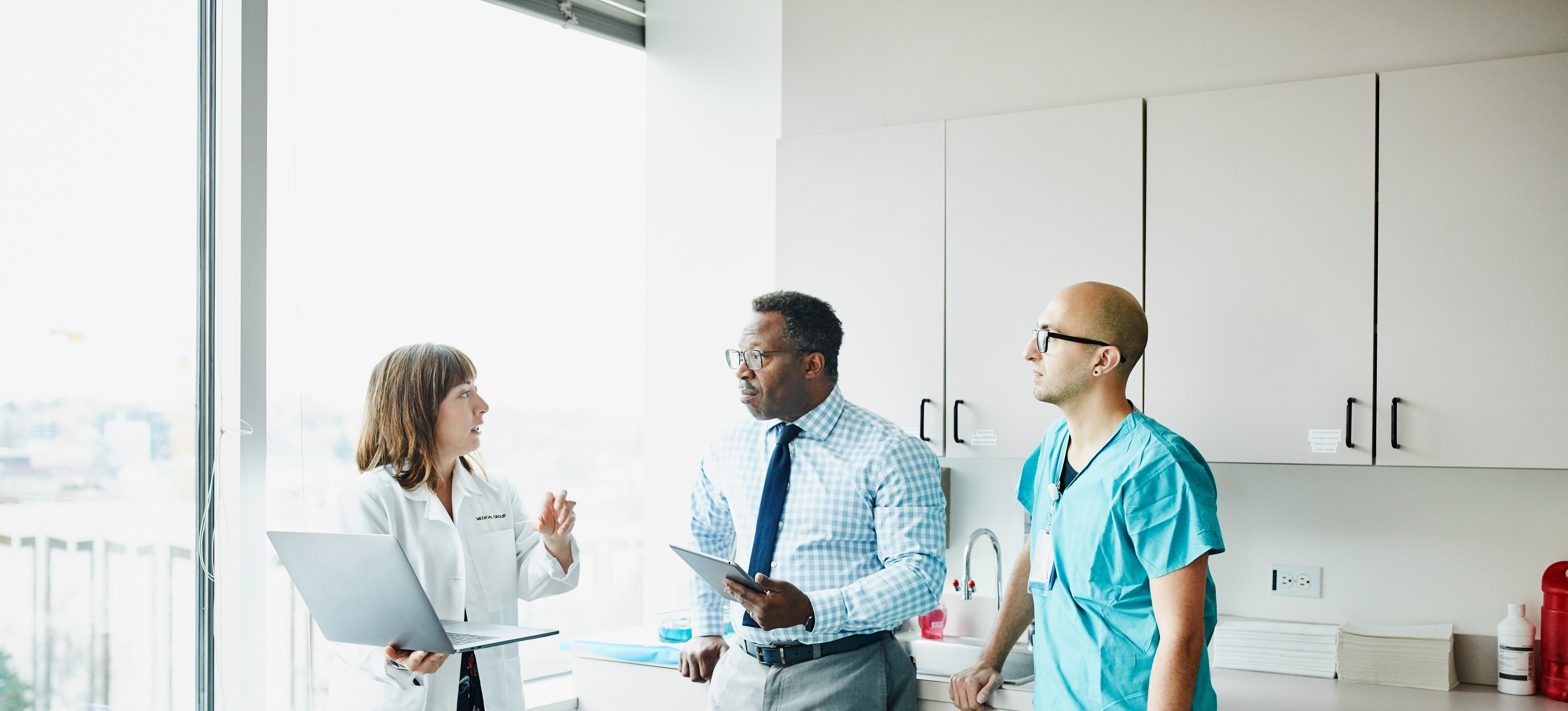 [Featured Image]:  A nursing home administrator, with short black hair, wearing glasses, wearing a blue white patterned shirt with a dark tie, is speaking to staff, one worker with a green uniform and one wearing a white coat, patterned dress, who is holding their laptop.
