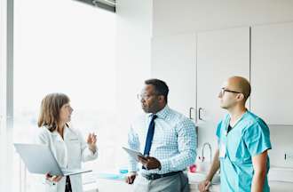 [Featured Image]:  A male, nursing home administrator, with short black hair, wearing glasses, wearing a blue white patterned shirt with a dark tie, is speaking to staff, one male with a green uniform and a female wearing a white coat, patterned dress, who is holding her laptop.