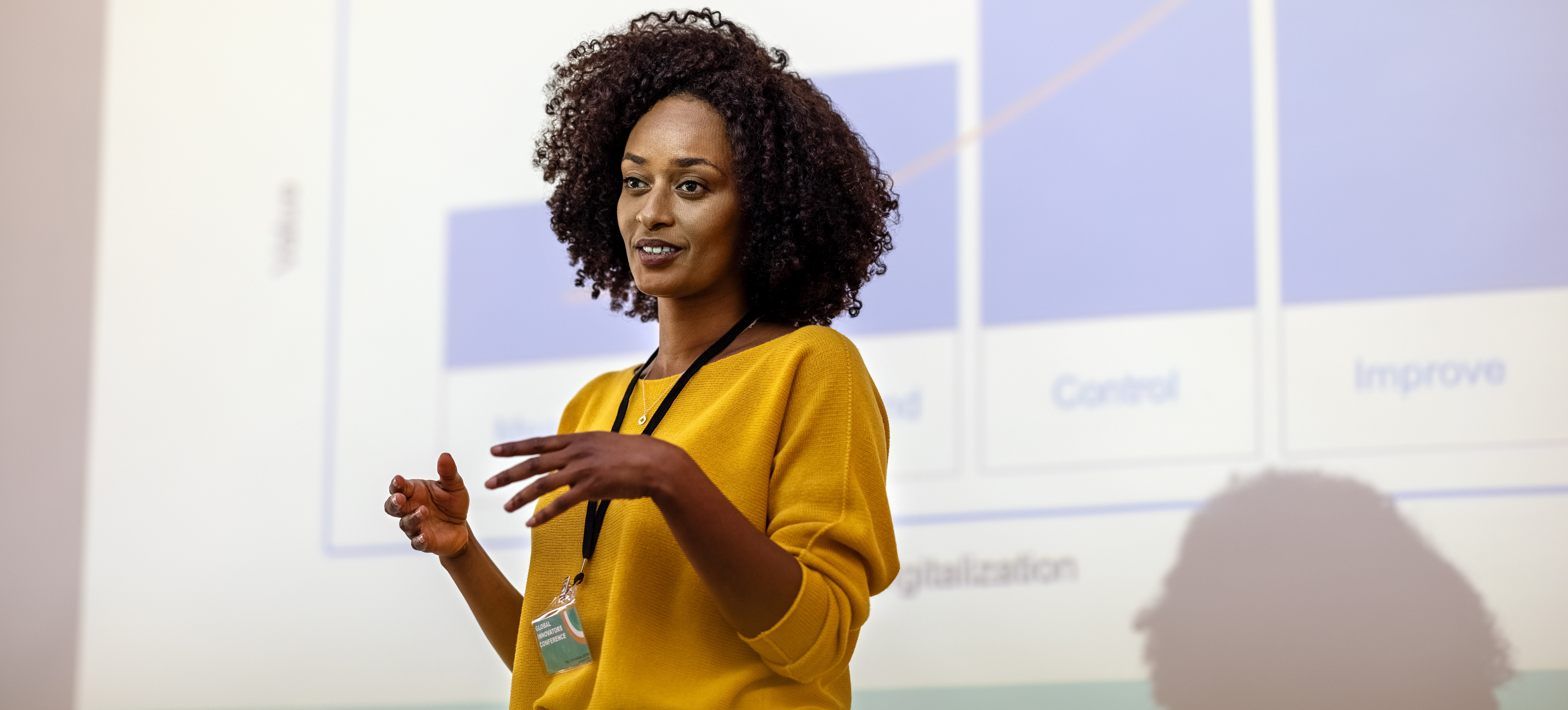 [Image en vedette] Une femme vêtue d'une chemise jaune fait une présentation d'affaires, y compris un rapport de données, avec un écran de projection avec des graphiques derrière elle.