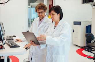 [Fetured imge] Two health information managers stand in a white room, looking at patient data on a laptop computer while standing in front of two desktop computers