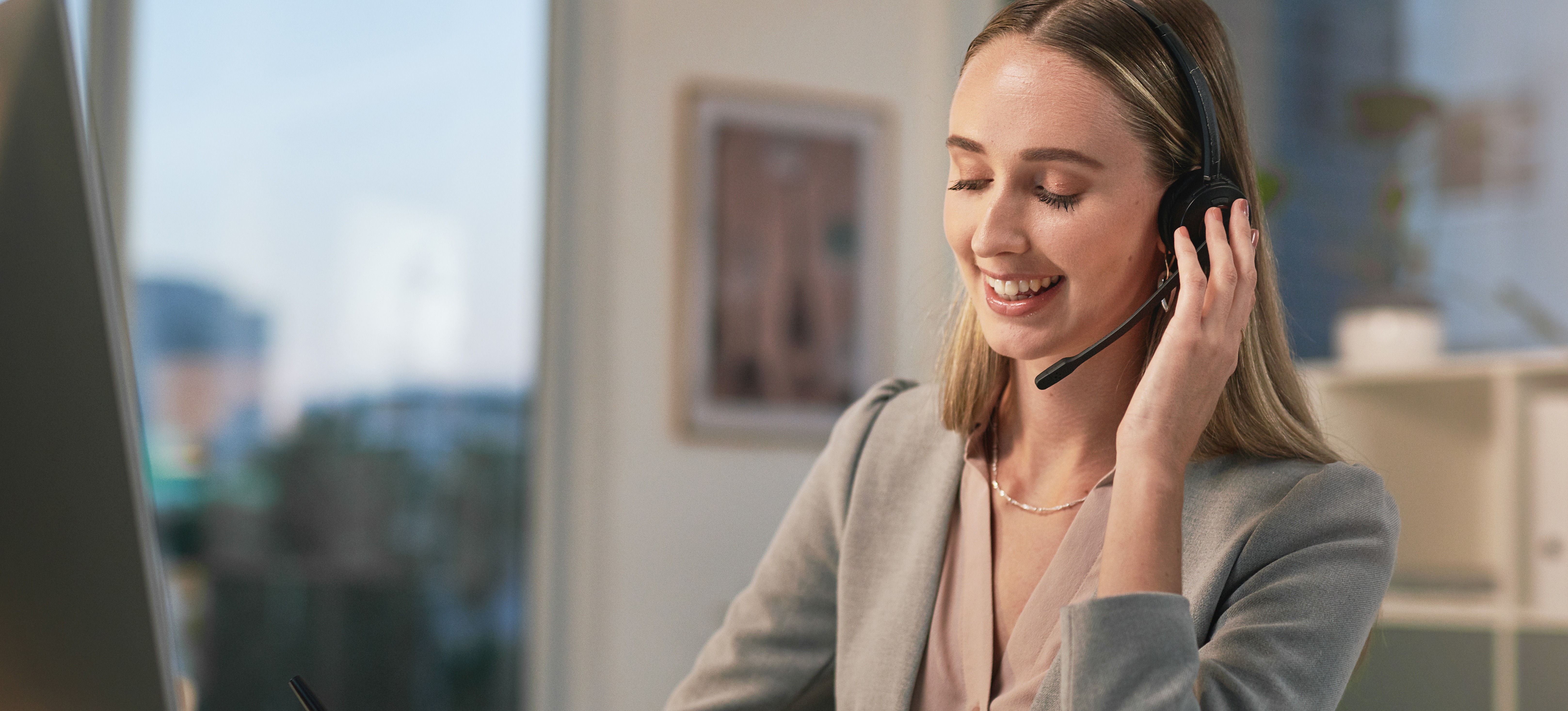 [Featured Image] A young woman working for an executive assistant salary sits at her desk in front of a computer, wearing a headset and taking notes.