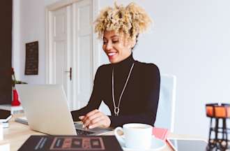 [Featured image] A person in a black turtleneck sweater sits at a table with books and a coffee cup and researches social media marketing certifications on their laptop.