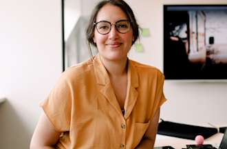 [Featured image] A UX designer in a yellow shirt and glasses sits at her computer, smiling at the camera.