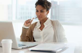 [Featured Image] A woman uses her laptop and paper to draft her SQL resume in an office setting. 