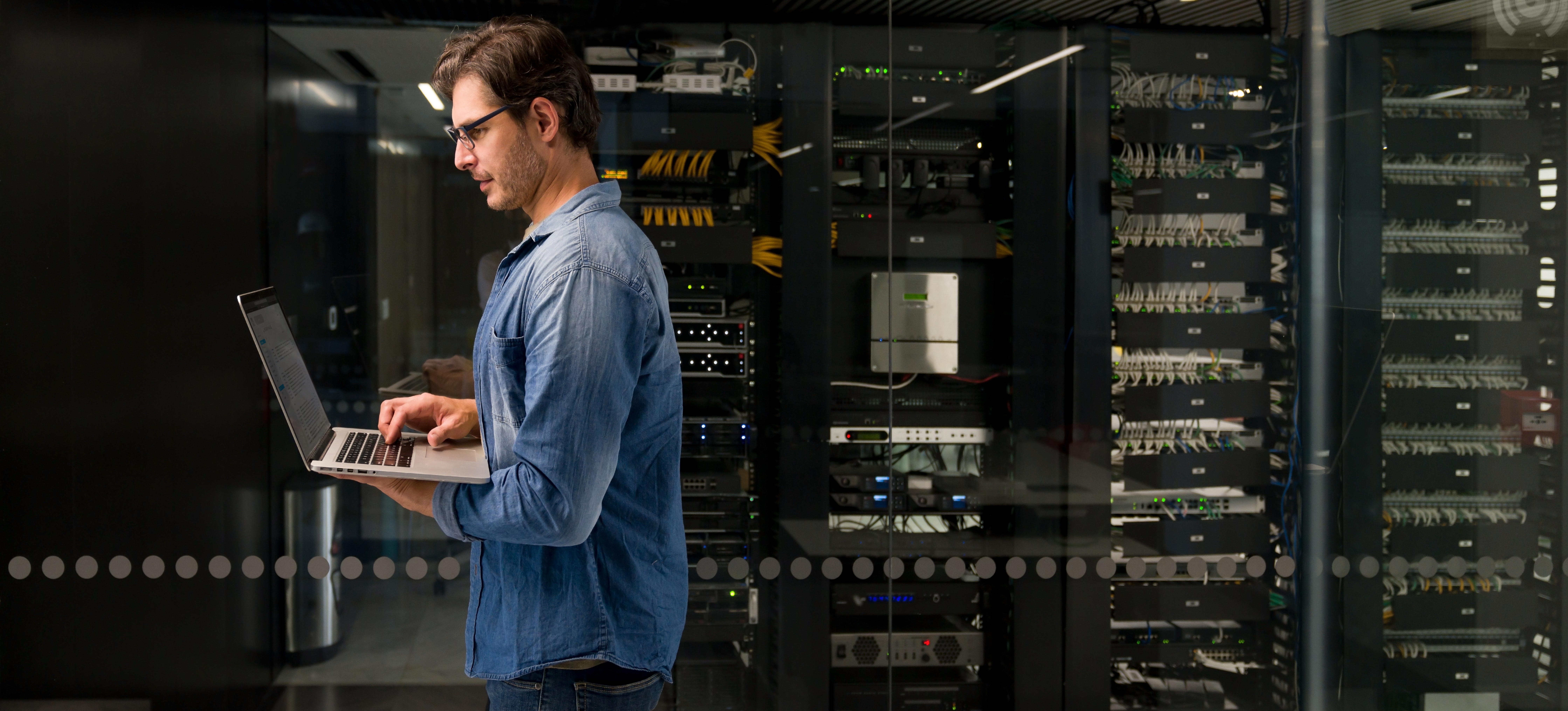[Featured Image] A data warehouse developer works on their laptop in a server room.
