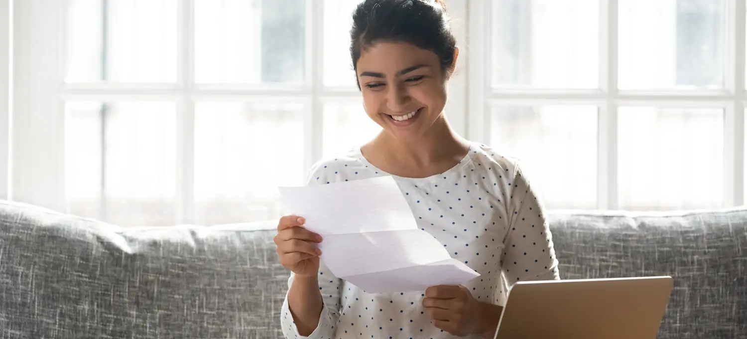 [Featured Image] A young, happy college graduate sitting in her living room reading their college transcripts.