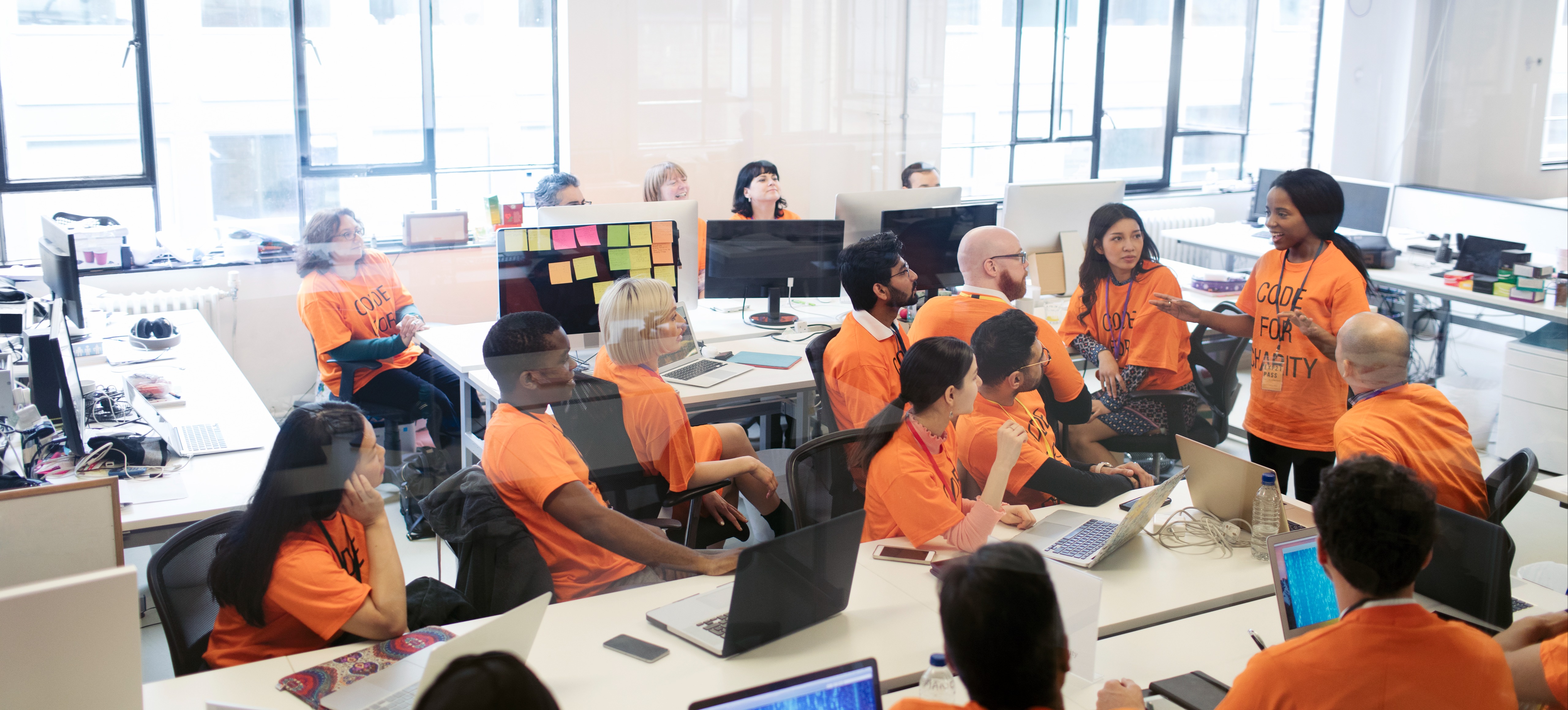 [Featured Image] After learning what a hackathon is, a group of young professionals participate in their first hackathon for charity, sitting at computers in a large office space, as one person addresses the group.
