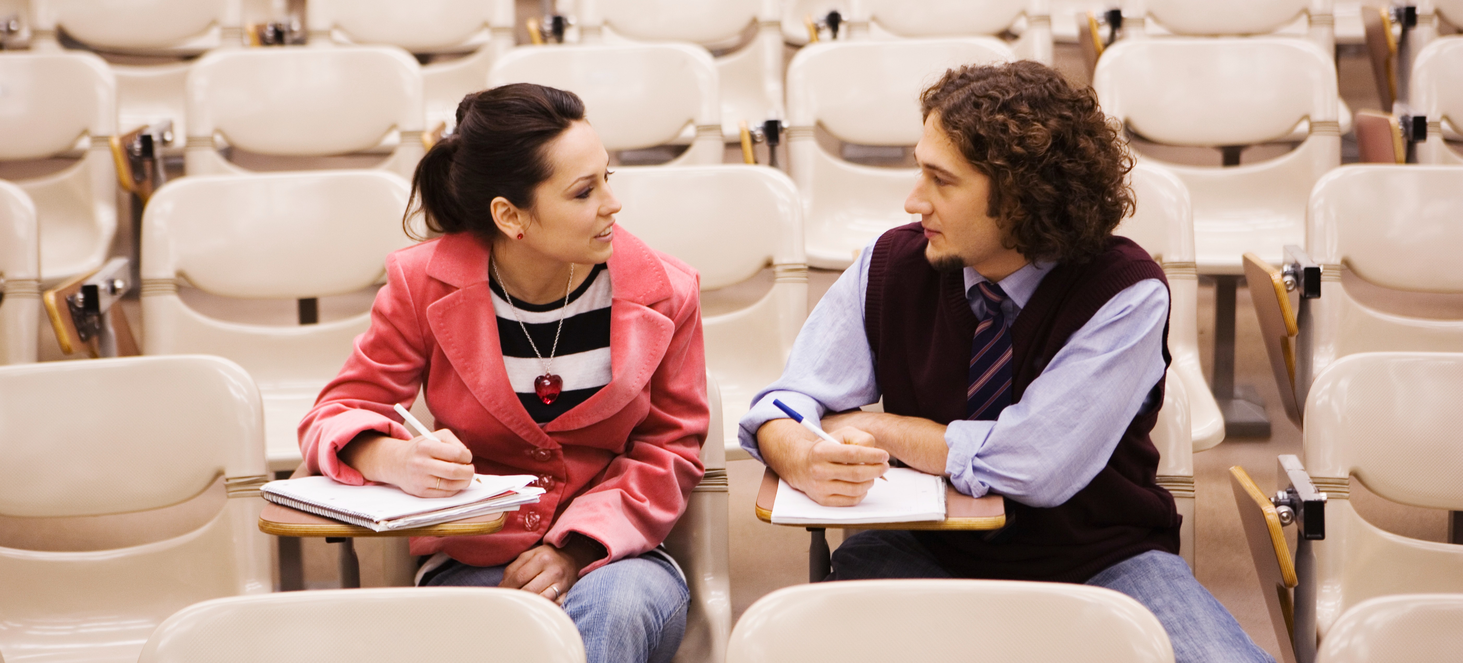 [Featured Image] Two students sit and talk in a college classroom, discussing what degree they need to become a lawyer.
