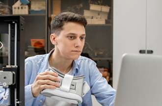 [Featured Image]:  Data Analyst, wearing a blue shirt, working on a laptop computer, and holding a headset, is working on technology skills. 