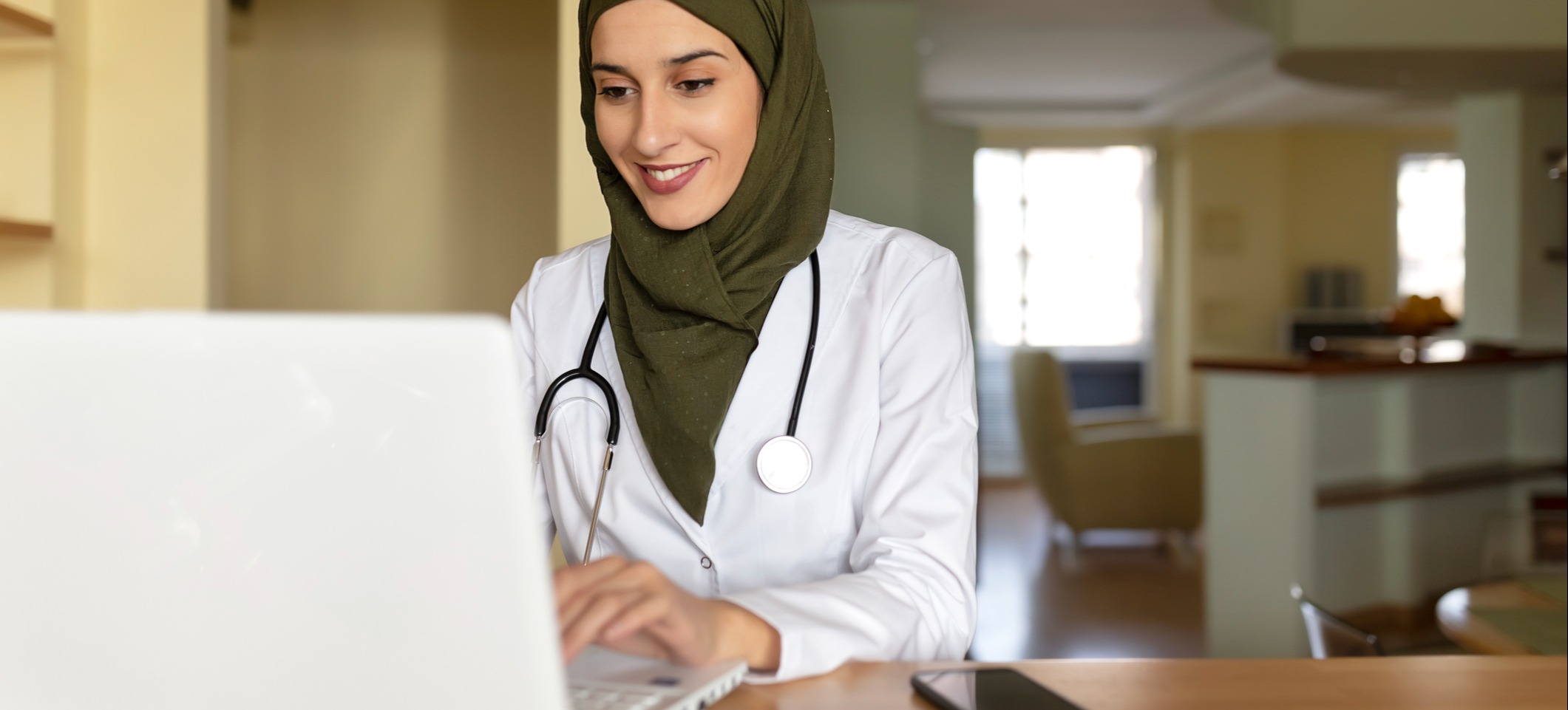 [Featured image] A DNP (Doctor of Nursing Practice) student in a lab coat, stethoscope, and head scarf works on their degree at a laptop computer.
