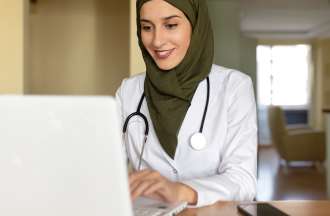 [Featured image] A DNP (Doctor of Nursing Practice) student in a lab coat, stethoscope, and head scarf works on her degree at a laptop computer.