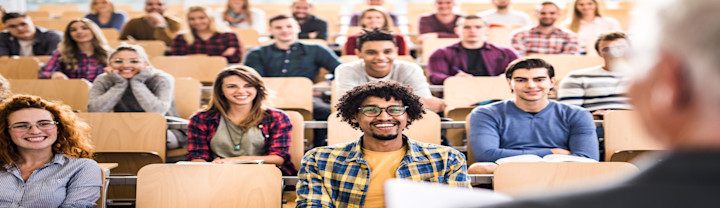 [Featured Image] A group of happy students deciding between trade school vs college sit at their desks, listening to their instructor at the front of the room.
