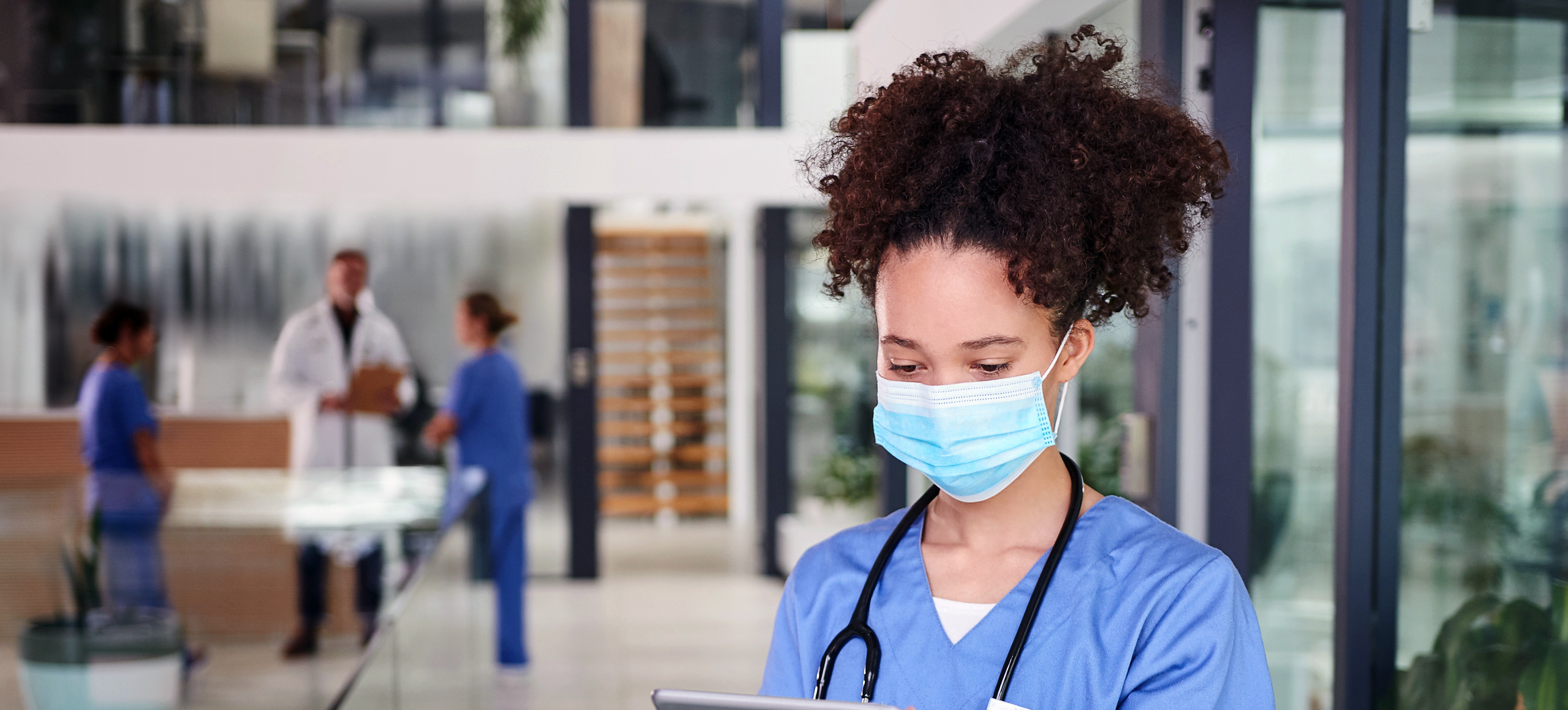 [Featured Image]: A worker with curly hair, wearing blue scrubs, a mask and a stethoscope around their neck is reading a chart. Doctors and nurses are in the background.