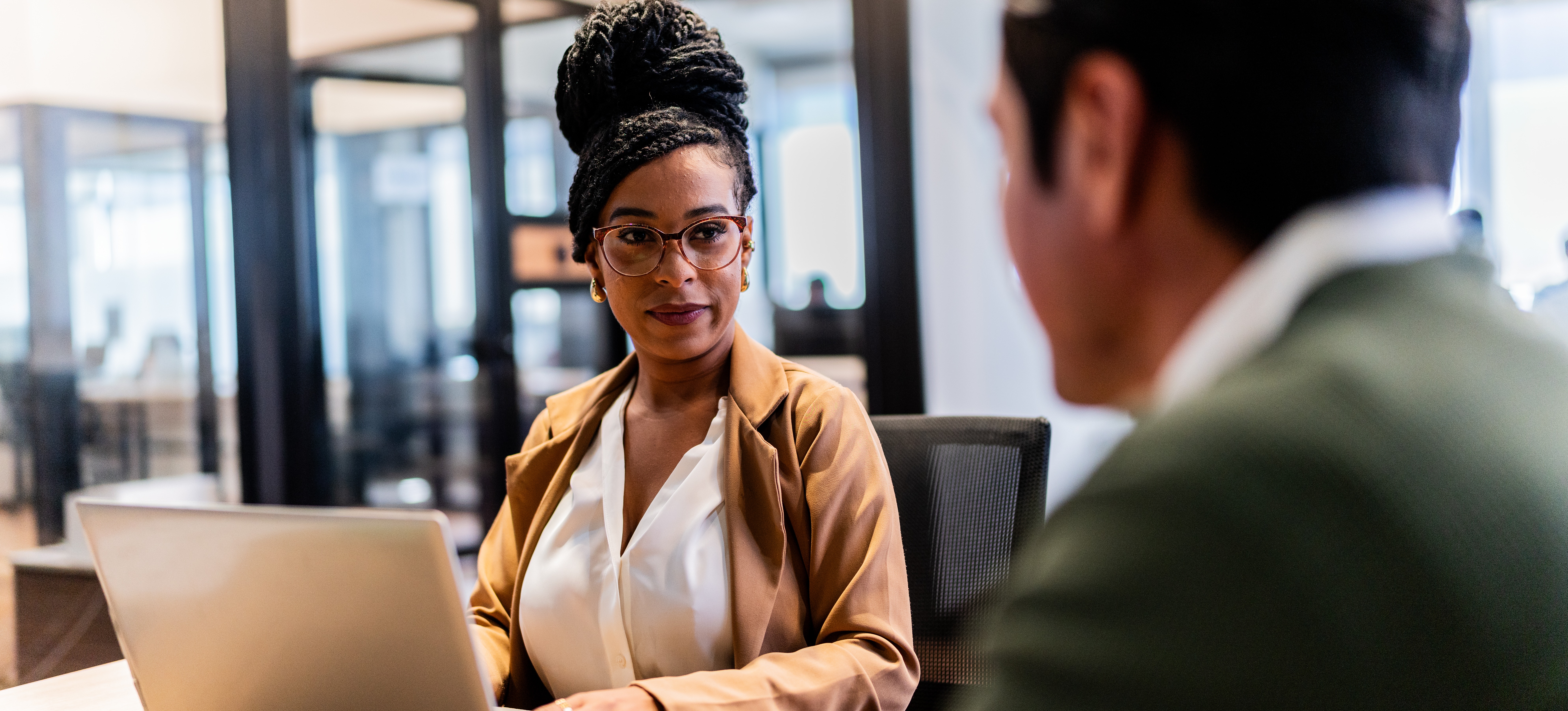 [Featured Image] A recruiter dressed in a white blouse and blazer listens to an interviewee answer their questions as they are poised to type notes on a laptop.
