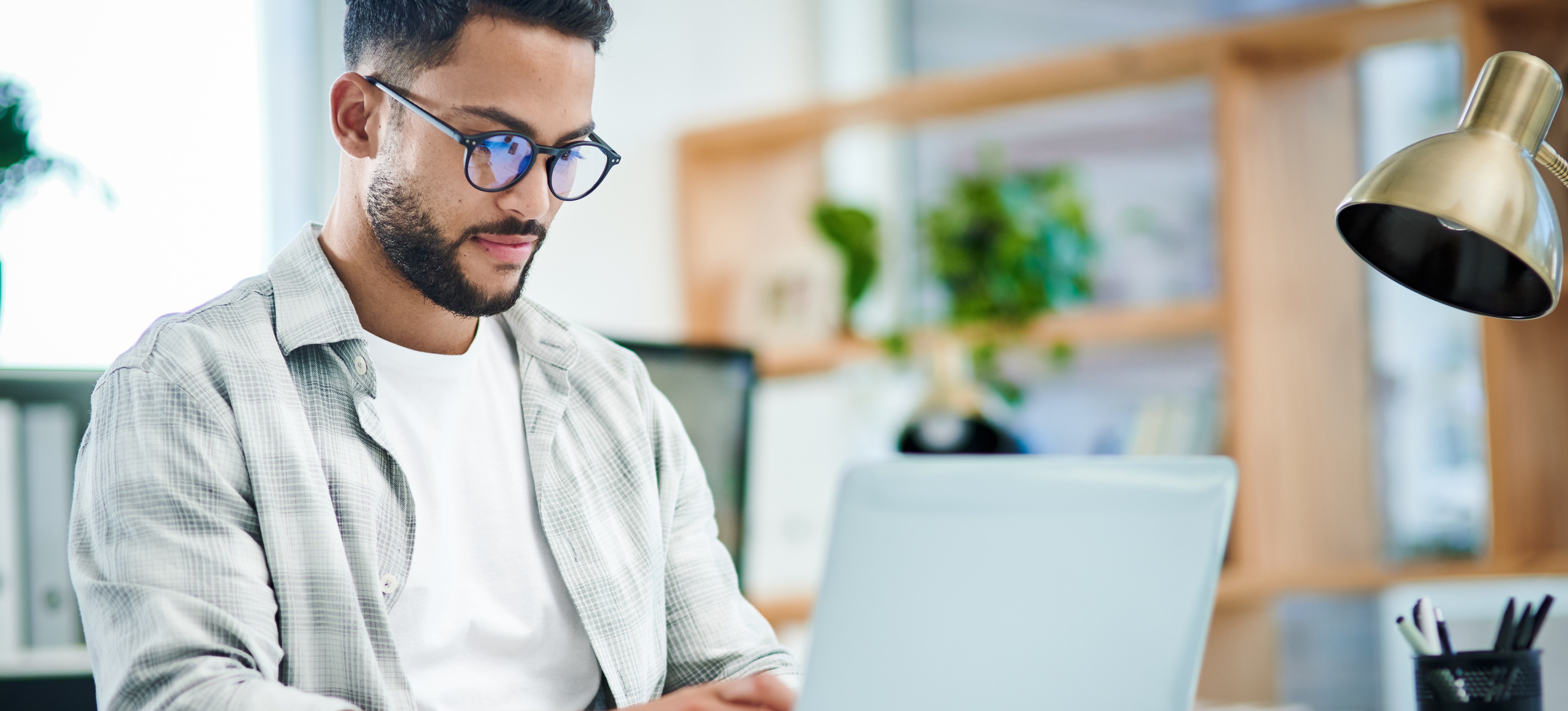 [Featured Image] A young businessman in a white shirt and wearing glasses works in an office on his laptop and learns how to create an NFT.
