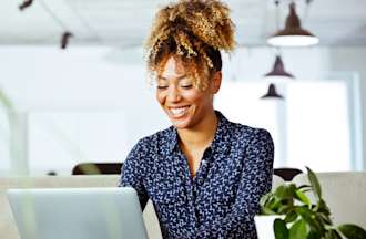 [Featured image] A young Black woman in a navy blue top smiles as she looks at her laptop.