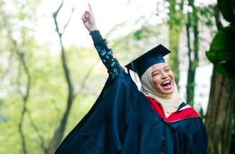 [Featured image] A first-generation college graduate wearing a cap and gown celebrates earning her bachelor's degree by throwing her hand in the air, holding up one finger.