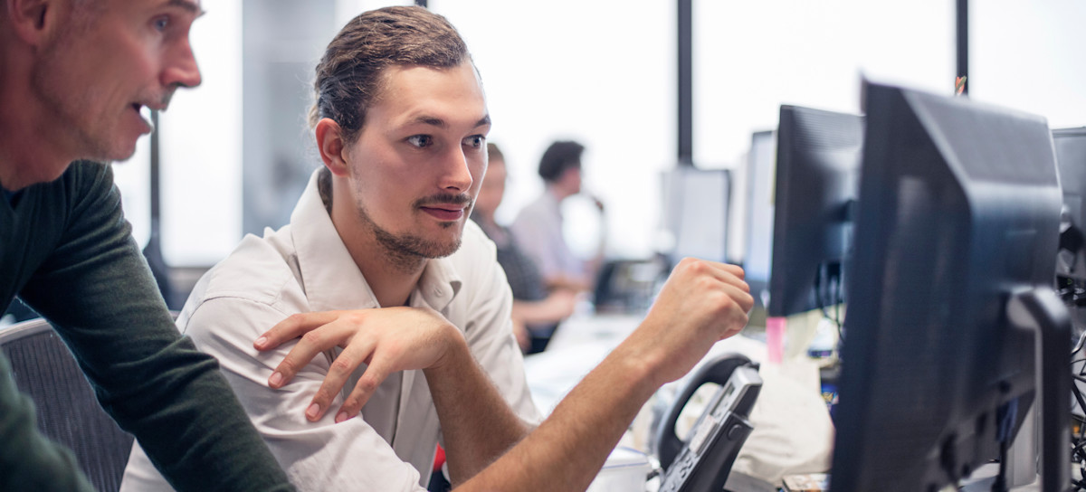 [Featured Image] Two blockchain developers look at a computer monitor while talking in an office setting with multiple workstations.

