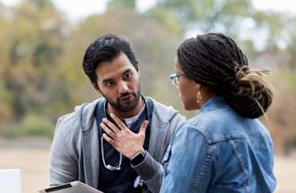 [Featured Image]:  A male care coordinator, wearing a gray sweatshirt, blue shirt, and a stethoscope around his neck, consultants with a patient as they sit on a park bench.