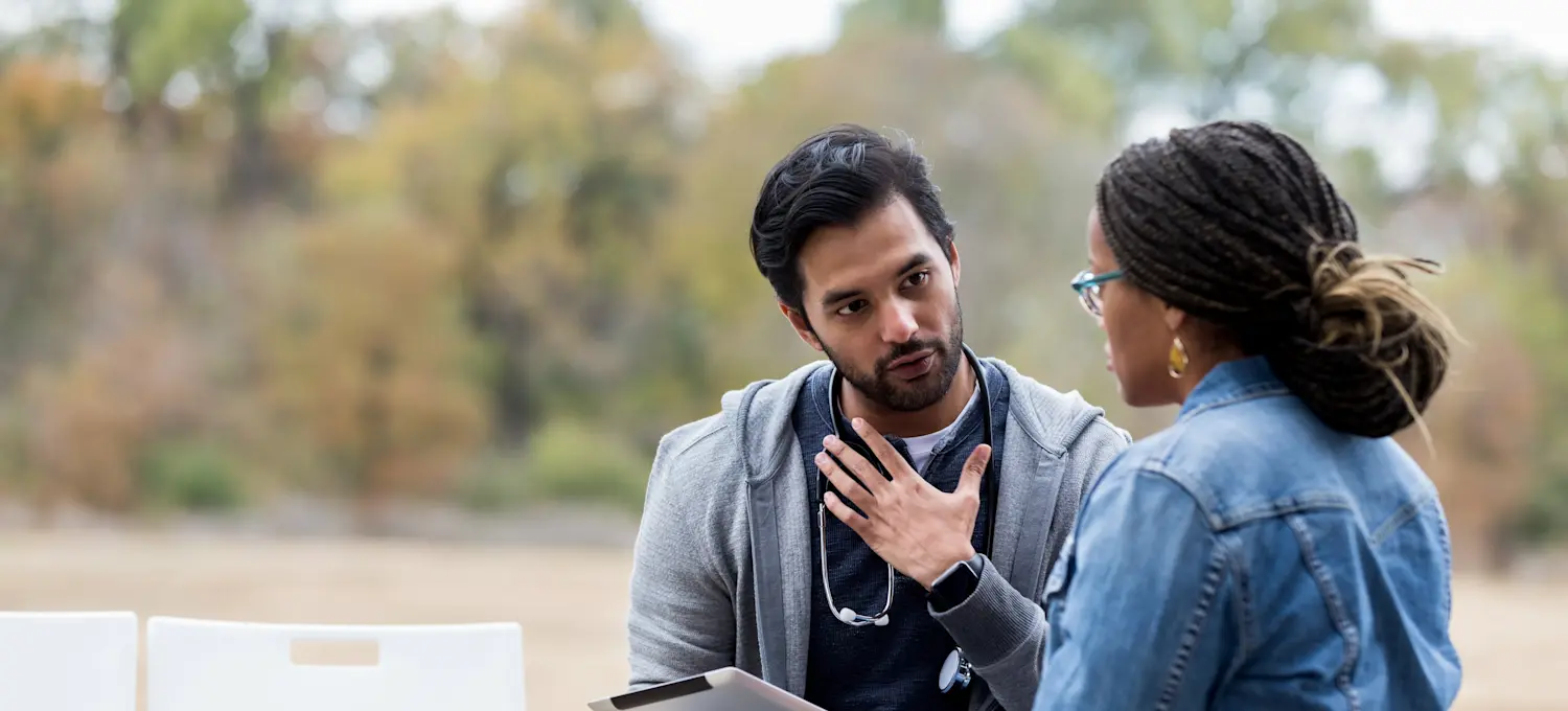 [Featured Image]:  A care coordinator, wearing a gray sweatshirt, blue shirt, and a stethoscope around their neck, consultants with a patient as they sit on a park bench.
