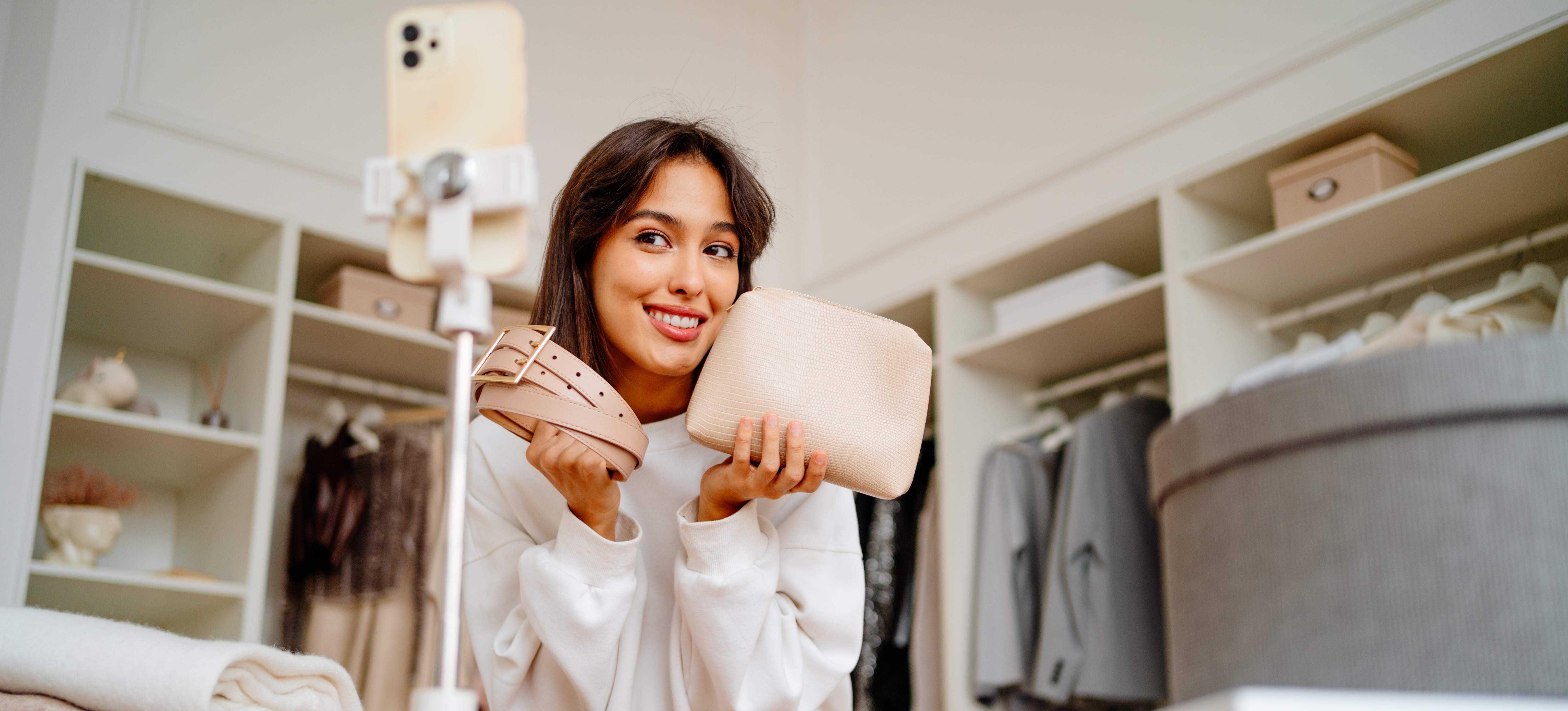 [Featured Image] Social influence at work: An influencer videos content on a smartphone, holding up a handbag and matching belt for the camera. 
