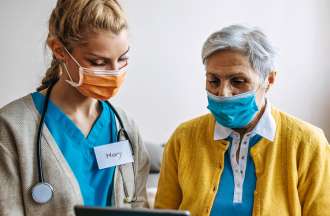 A younger nurse wearing a face mask, blue scrubs, and stethoscope assists an older women in a yellow sweater