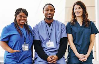 Three nurses, two female and one male all wearing blue scrubs, smile in a hospital. 