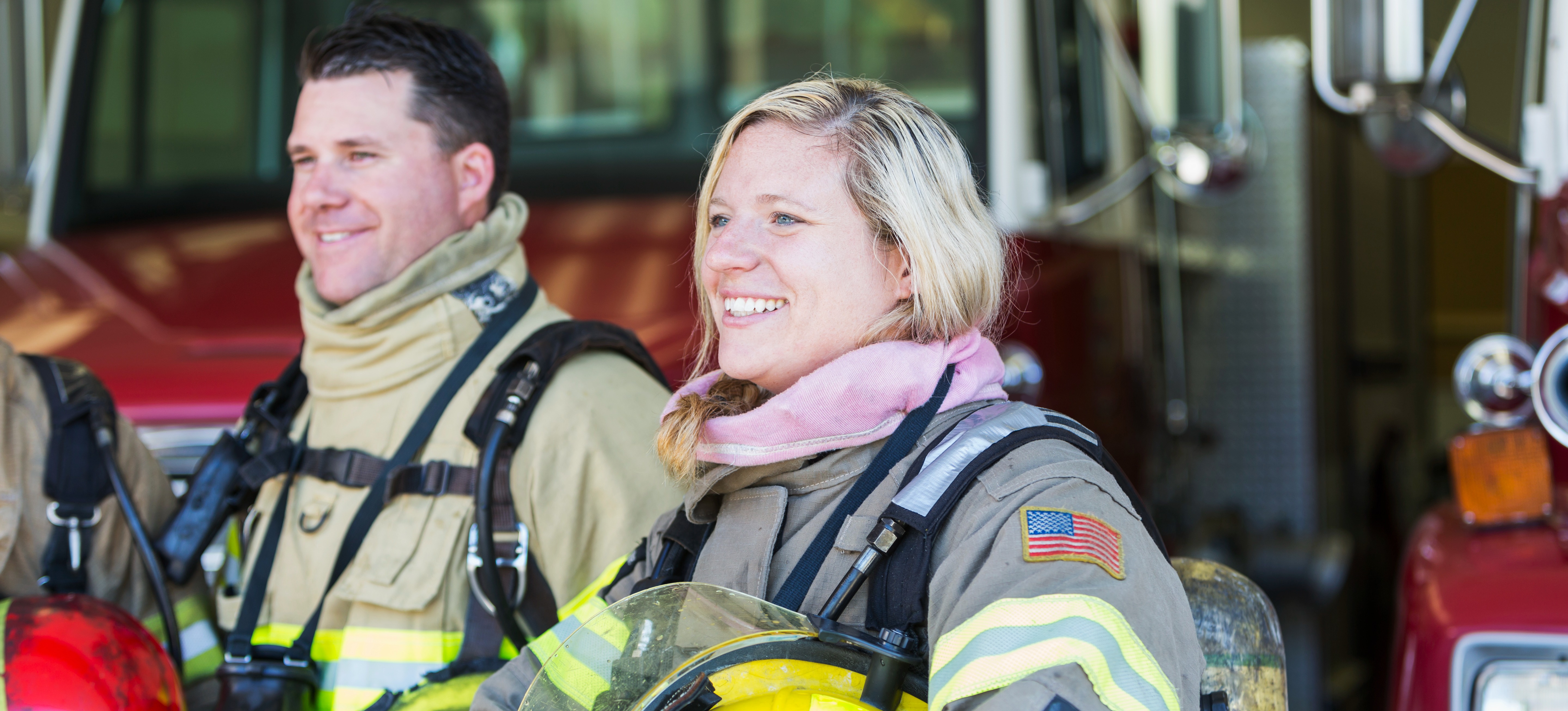 [Featured Image] Two smiling first responders dressed in clothing to fight fires look off into the distance while standing in front of fire trucks.
