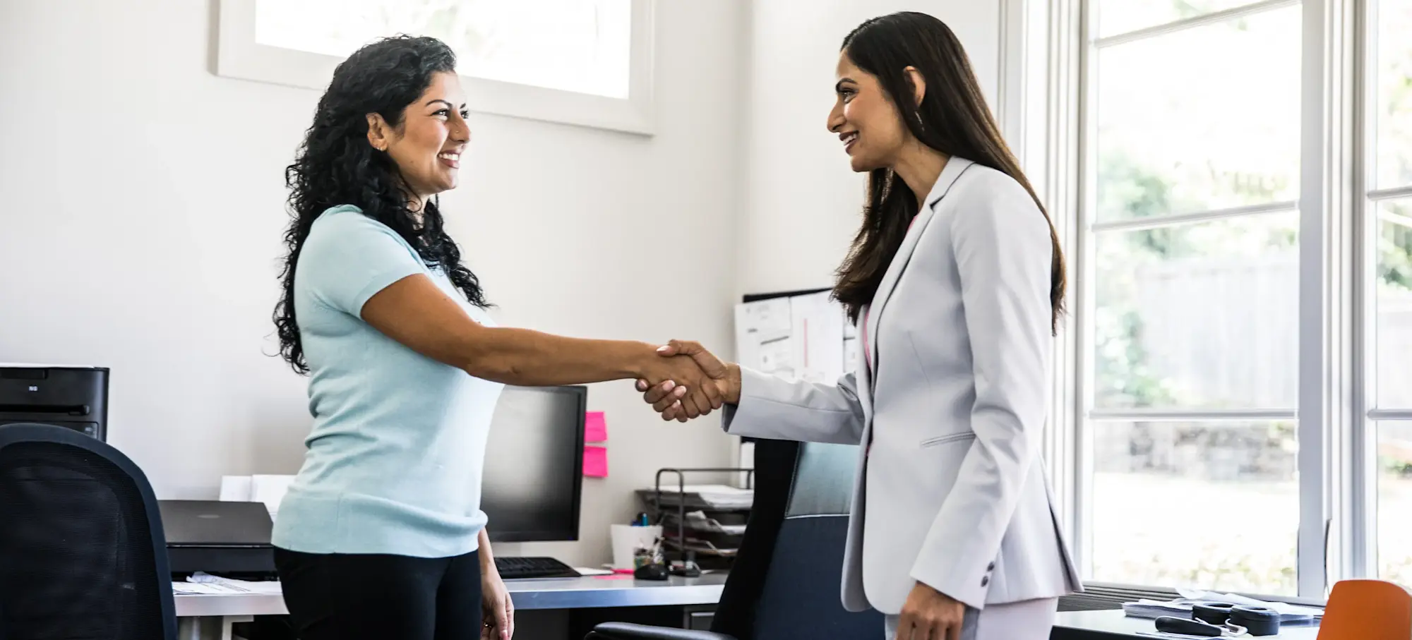 [Featured Image] Two women are shaking hands as they meet in an office for an IT interview.