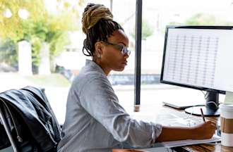 [Featured image] A woman in glasses and a striped shirt sits in front of a compute searching for business degree jobs while writing on a notepad.