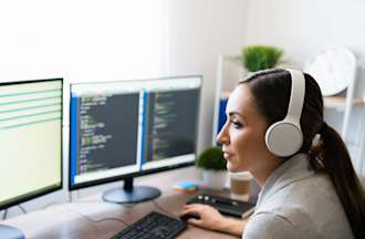 [Featured Image] A female, wearing a gray top and white headphones, sits in front of two desktop computers.