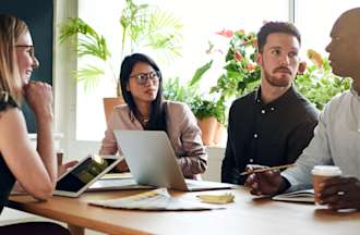 [Featured image] A marketing team sits at a wooden table discussing content strategy. Some are working on tablets, laptops, and notebooks.