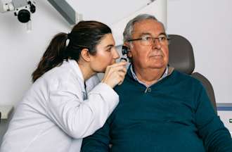 [Featured Image] A female audiologist examines a male patient in a clinical setting. 