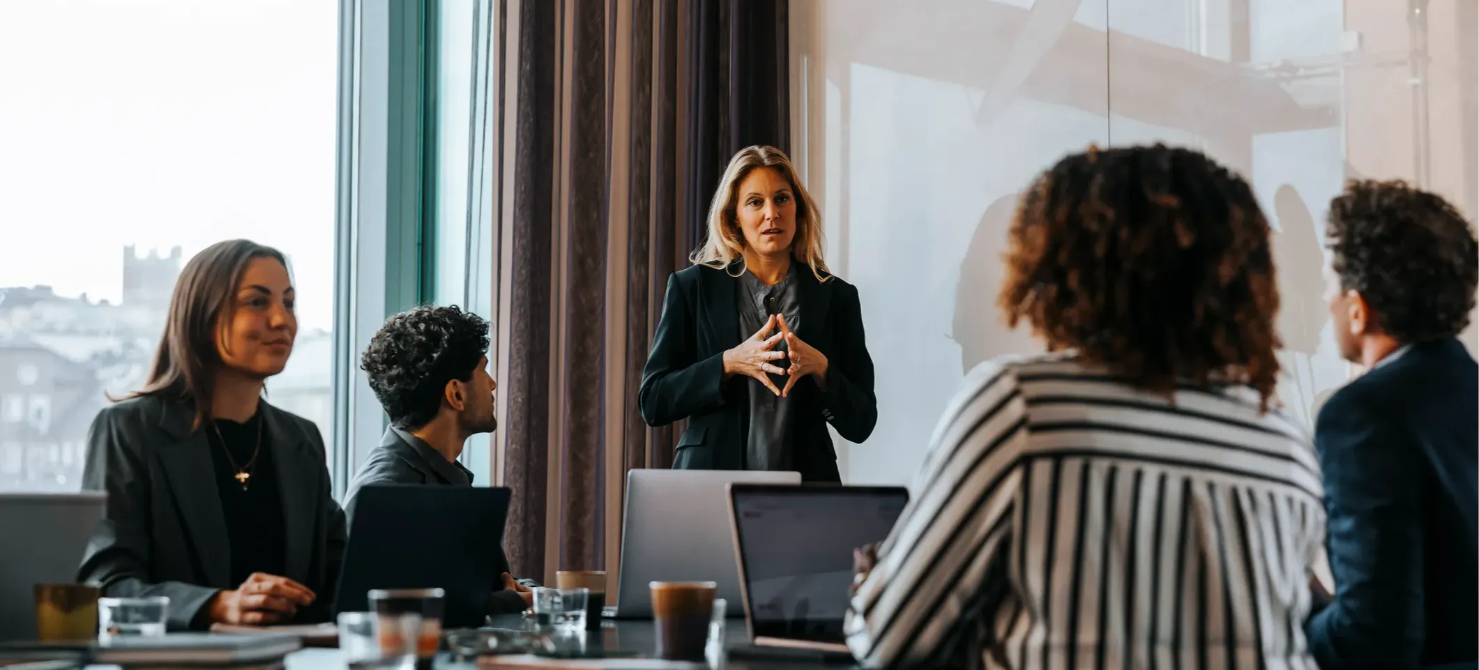 [Featured image]: A person stands in a boardroom as they presents a corporate finance report to colleagues sitting around the table.
