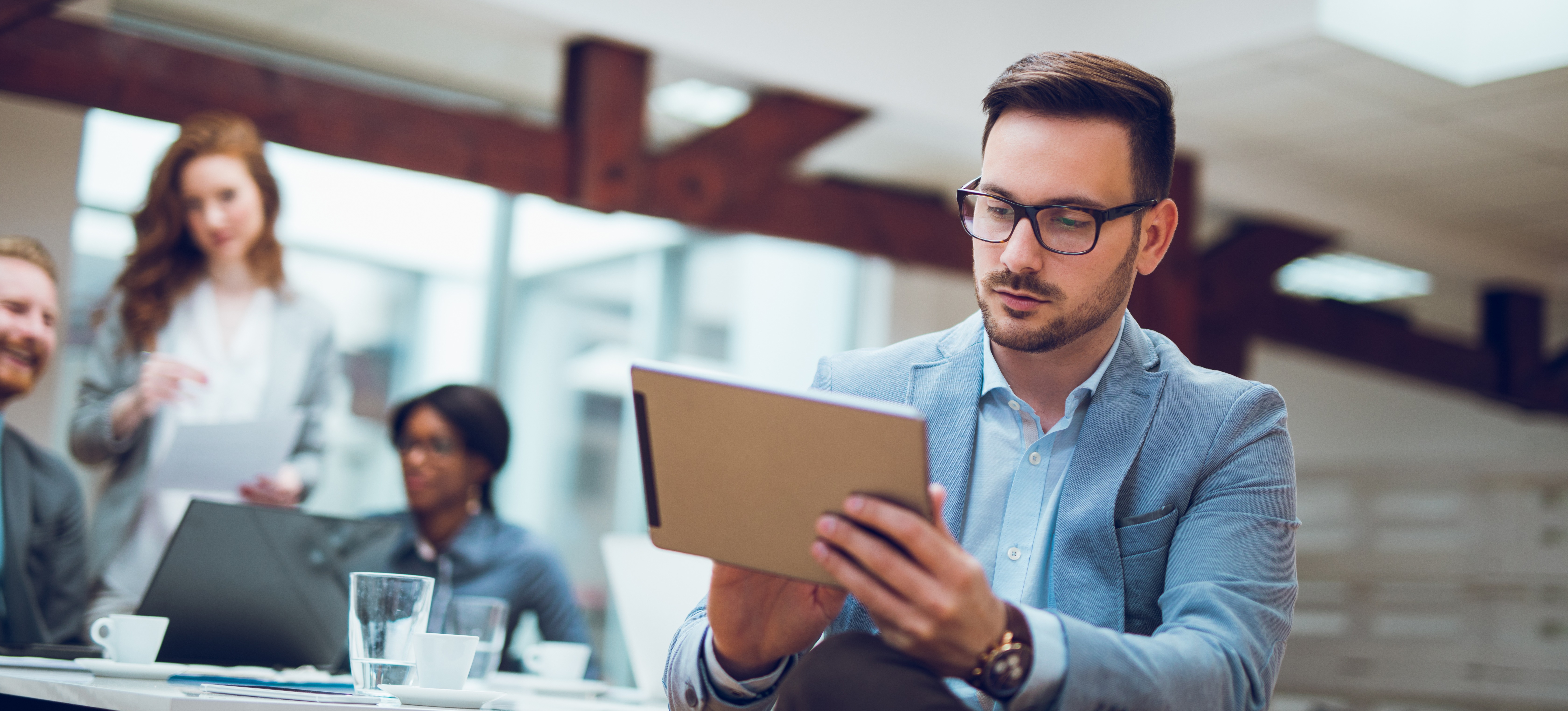 [Hauptbild] Person, die an einem Tablet arbeitet, während Kollegen eine Diskussion führen. 