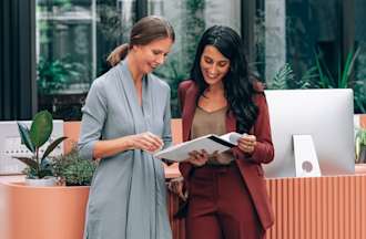 [Featured Image] An assistant project manager and a project manager stand in an office, reviewing notes from a project meeting.