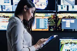 [Featured image] A cybersecurity tech in a gray shirt stands up looking at a tablet in front of a wall of video security monitors.