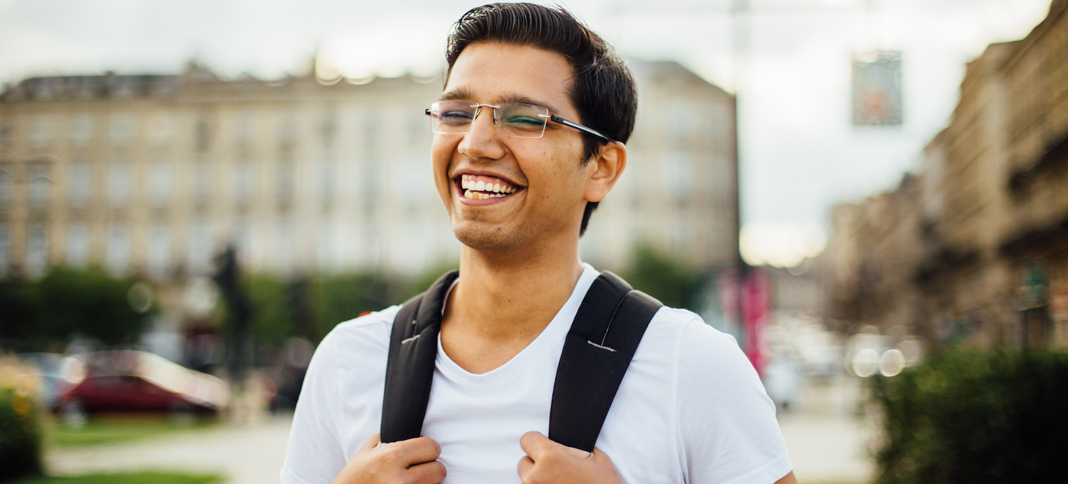 [Featured image] A young learner in glasses and a light blue shirt stands smiling and gripping the straps of their backpack. 
