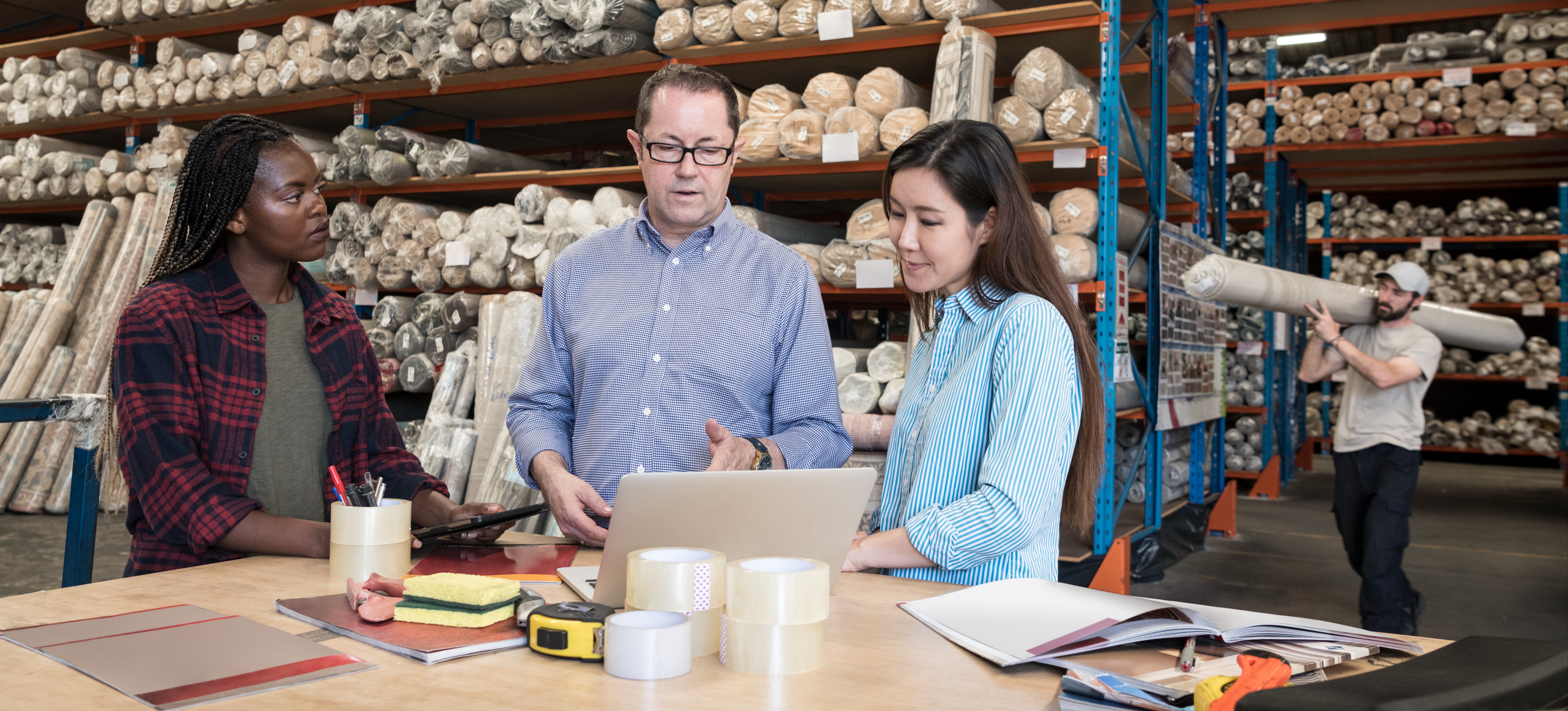 [Featured Image] A business analyst discusses revenue optimization with colleagues while looking at a laptop and standing in a carpet warehouse.
